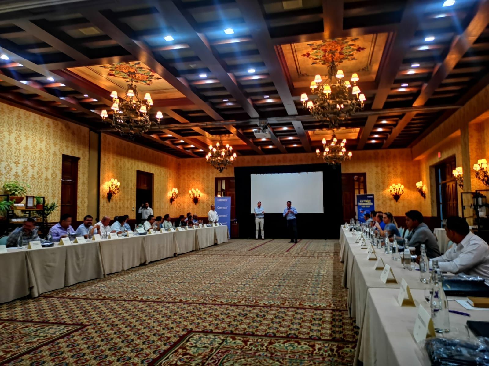 Salón de la reunión mensual de AGIM Los Cabos en mayo de 2023, con mesas en herradura y pantalla de presentación